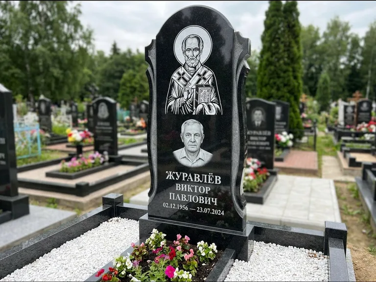 Памятник с гравировкой иконы: стела с образом в полный рост на обороте — гравировка образа Матроны Московской, низкие кусты спиреи в цвету, работа «Лебедева» в Ханты-Мансийске