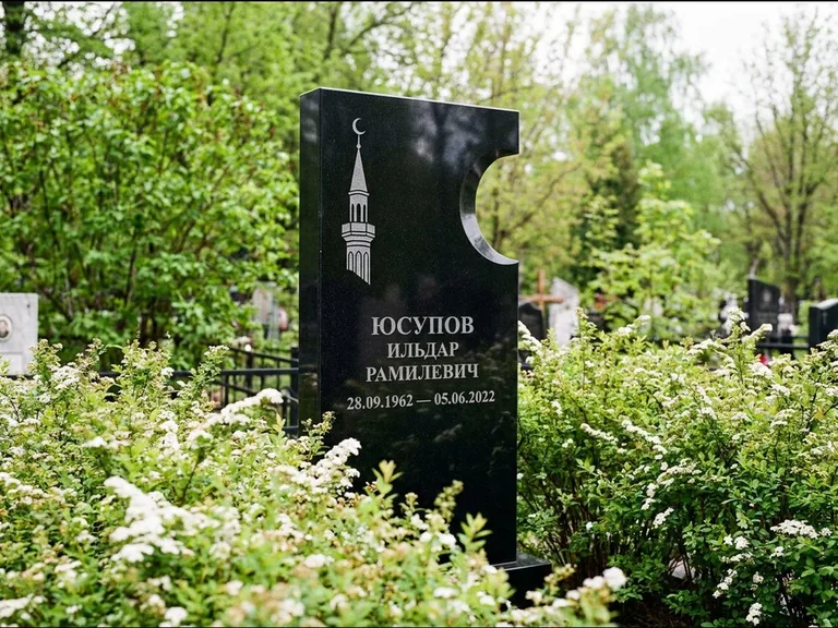 Мусульманский памятник с гравировкой мечети: стела с боковым вырезом в форме полумесяца — гравировка минарета с полумесяцем, низкие кусты спиреи в цвету, работа «Юсупов» в Ханты-Мансийске